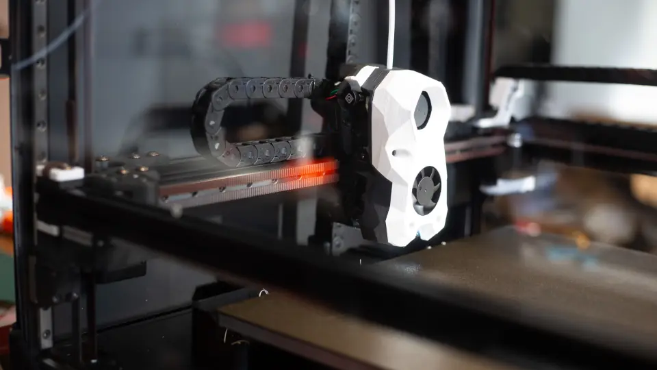 The print head of a desktop 3D printer on its rail, the drag chain behind it and an empty build plate below.